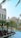 A scenic urban waterfront featuring a row of palm trees lining a canal with clear blue water. Modern Middle Eastern architecture is showcased in the buildings, with intricate designs and beige facades. Seated people are visible at outdoor cafes, adding a leisurely vibe.