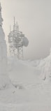 A telecommunications tower heavily covered in snow and ice, situated in a snowy landscape. The surrounding area is blanketed with thick snow, giving an isolated, wintery appearance. The sky is overcast, adding to the cold and serene atmosphere.