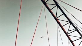 Red structural elements of a bridge or tower against a pale sky, with an airplane flying in the background.