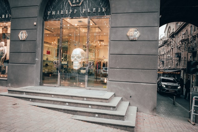 A store entrance featuring a large glass door with a prominent skull artwork displayed inside. The building is dark-colored with glossy finishes, and there are steps leading up to the entrance. The surrounding area includes city street elements like cars and other buildings.