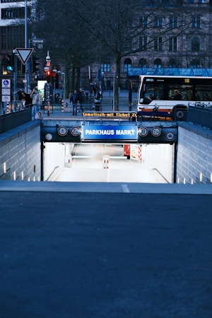 A city street scene features a bus stopped near the entrance to an underground parking garage marked 'PARKHAUS MARKT'. Trees without leaves and older-style buildings can be seen in the background. People are walking nearby, and there's a bicycle parked at the fence.