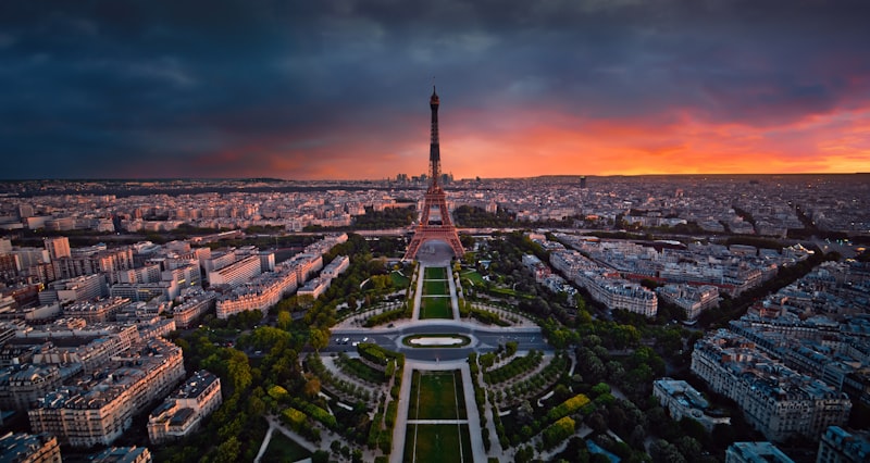Aerial view of the Eiffel Tower and the tree-lined Champ de Mars at sunset, with the Paris skyline under dramatic clouds