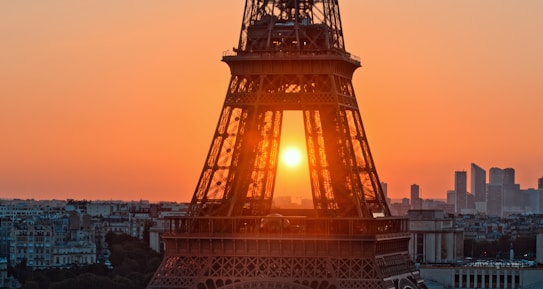The image features the Eiffel Tower during sunset. The sun is positioned directly behind the structure, creating a silhouette effect. The sky is a gradient of warm hues transitioning from deep orange to a softer yellow. In the background, the skyline of Paris is visible with various buildings.
