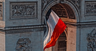 A large French flag billows beneath the arch of a historical monument adorned with intricate stone carvings and sculptures. The architecture displays a combination of reliefs and ornate designs, showcasing classical art.