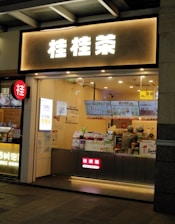 A small bubble tea shop with illuminated signage in front. Inside, two staff members are wearing uniforms and preparing orders. Various promotional posters and a digital menu displaying drink options are visible on the counter.
