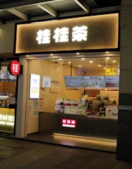 A small bubble tea shop with illuminated signage in front. Inside, two staff members are wearing uniforms and preparing orders. Various promotional posters and a digital menu displaying drink options are visible on the counter.