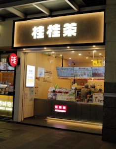 A small bubble tea shop with illuminated signage in front. Inside, two staff members are wearing uniforms and preparing orders. Various promotional posters and a digital menu displaying drink options are visible on the counter.
