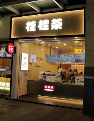 A small bubble tea shop with illuminated signage in front. Inside, two staff members are wearing uniforms and preparing orders. Various promotional posters and a digital menu displaying drink options are visible on the counter.