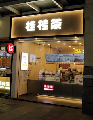 A small bubble tea shop with illuminated signage in front. Inside, two staff members are wearing uniforms and preparing orders. Various promotional posters and a digital menu displaying drink options are visible on the counter.