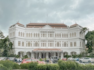 A grand white colonial-style building with multiple arched windows and a central entrance. The facade is adorned with decorative moldings and a rooftop with red tiles. Several cars are parked in front of the building, surrounded by lush greenery, and a few people are walking near the entrance.