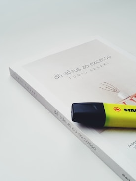 A book titled 'd&ecirc; adeus ao excesso' by Fumio Sasaki lies on a light background. The book cover is minimalistic with soft colors, featuring a simple graphic. A yellow and black highlighter is placed on top of the book, positioned at an angle.
