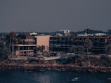 A modern building complex is located near the waterfront surrounded by tall palm trees and vegetation. The structure features multiple levels with glass windows and balconies. In the background, a dense line of foliage and other urban structures are visible under a clear sky.