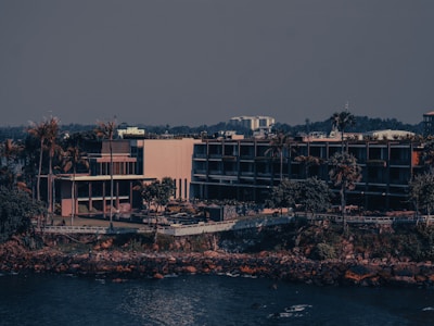 A modern building complex is located near the waterfront surrounded by tall palm trees and vegetation. The structure features multiple levels with glass windows and balconies. In the background, a dense line of foliage and other urban structures are visible under a clear sky.