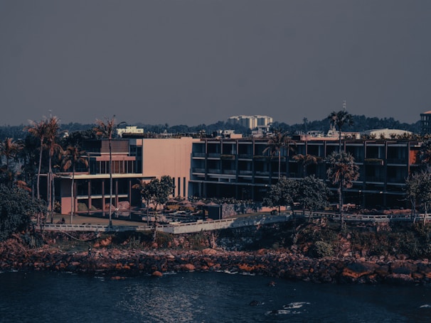 A modern building complex is located near the waterfront surrounded by tall palm trees and vegetation. The structure features multiple levels with glass windows and balconies. In the background, a dense line of foliage and other urban structures are visible under a clear sky.