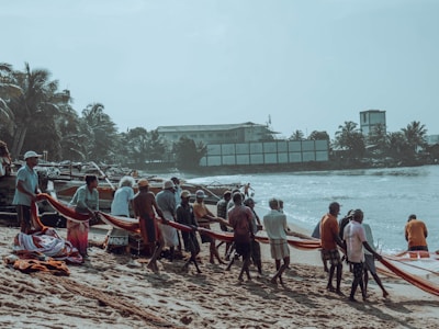 A group of people on a sandy beach are collaboratively pulling in a fishing net from the sea. The setting is tropical, with palm trees and a few buildings visible in the background.
