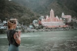 A person with tattoos is standing by a riverside with hands in a prayer position, overlooking a large, ornate temple complex situated among lush green hills. The architecture of the temple features multiple spires and intricate detailing, creating a serene and contemplative atmosphere.