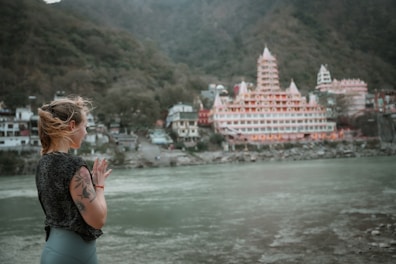 A person with tattoos is standing by a riverside with hands in a prayer position, overlooking a large, ornate temple complex situated among lush green hills. The architecture of the temple features multiple spires and intricate detailing, creating a serene and contemplative atmosphere.