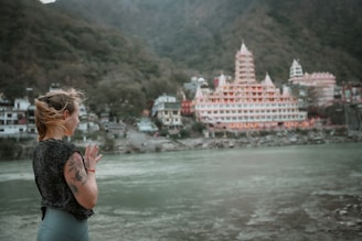 A person with tattoos is standing by a riverside with hands in a prayer position, overlooking a large, ornate temple complex situated among lush green hills. The architecture of the temple features multiple spires and intricate detailing, creating a serene and contemplative atmosphere.