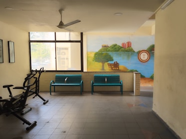 A room with exercise equipment on one side, including stationary bikes, and two blue benches against a wall. A large window provides natural light, and a bright mural adorns the wall, depicting a serene lakeside scene with greenery and a couple sitting on a bench.