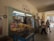A small convenience store with shelves stocked with various products including canned goods, drinks, and snacks. The entrance is open with fluorescent lighting inside. Two people are interacting outside the store, and there is an ice cream freezer prominently displayed.