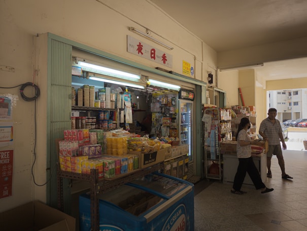 A small convenience store with shelves stocked with various products including canned goods, drinks, and snacks. The entrance is open with fluorescent lighting inside. Two people are interacting outside the store, and there is an ice cream freezer prominently displayed.