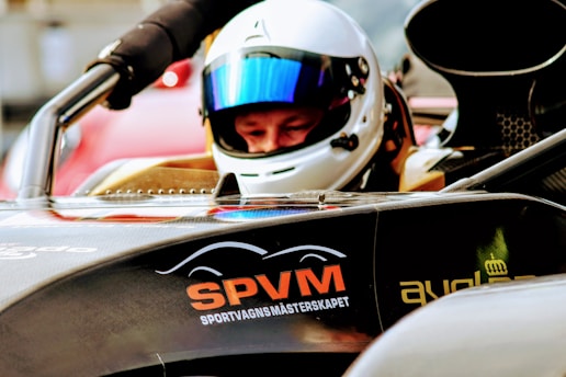 A person wearing a white helmet and reflective visor sits inside a racing car. The front of the car displays the letters 'SPVM' in orange and additional text beneath. The car has a sleek, aerodynamic design with visible components and a dark color. The background is blurred, highlighting the focus on the driver and vehicle.