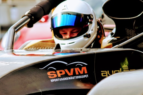 A person wearing a white helmet and reflective visor sits inside a racing car. The front of the car displays the letters 'SPVM' in orange and additional text beneath. The car has a sleek, aerodynamic design with visible components and a dark color. The background is blurred, highlighting the focus on the driver and vehicle.