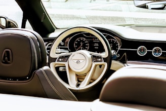 Interior view of a luxury car, focusing on the beige and black steering wheel with the manufacturer's logo prominently displayed. The dashboard features a digital display with various controls and analog gauges. The seats have elegant stitching, and the overall design exudes sophistication.