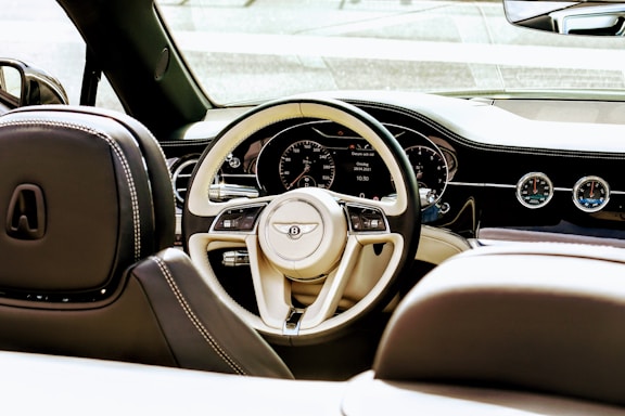 Interior view of a luxury car, focusing on the beige and black steering wheel with the manufacturer's logo prominently displayed. The dashboard features a digital display with various controls and analog gauges. The seats have elegant stitching, and the overall design exudes sophistication.