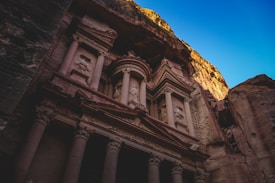 An ancient archaeological site features an intricately carved rock facade, with large columns and detailed stone reliefs. The structure is asymmetrically hewn into a massive stone cliff under a clear blue sky, illuminated by warm, golden sunlight that casts shadows enhancing its intricate details.