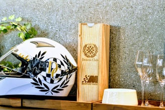 A white racing helmet adorned with black laurel designs and a checkered pattern sits on a wooden shelf. Beside it is a wooden box with the engraving '2022 Drivers Club' and 'Mantorp Park' logos. There are also empty wine glasses and a white ceramic dish nearby. Green foliage peeks in from the left side.