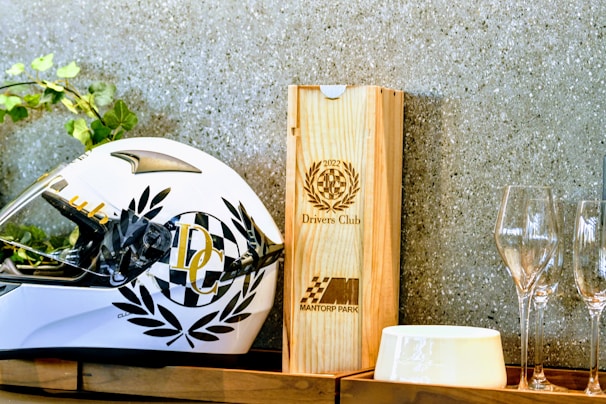 A white racing helmet adorned with black laurel designs and a checkered pattern sits on a wooden shelf. Beside it is a wooden box with the engraving '2022 Drivers Club' and 'Mantorp Park' logos. There are also empty wine glasses and a white ceramic dish nearby. Green foliage peeks in from the left side.