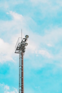 A tall, red-brown metallic tower with spotlights on top stands against a bright blue sky with scattered white clouds. The structure is industrial in design with a lattice platform beneath the lights.