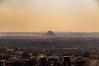 A vast urban landscape stretches beneath a hazy sky with the iconic pyramids visible in the background, standing majestically amidst the distant desert horizon.