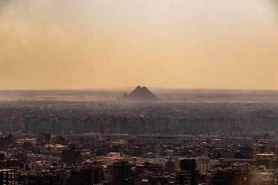 A vast urban landscape stretches beneath a hazy sky with the iconic pyramids visible in the background, standing majestically amidst the distant desert horizon.