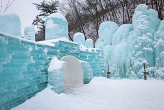 Intricate ice structures resembling castle walls are formed with large, smooth blocks of blue ice. The scene includes natural ice formations resembling frozen waterfalls against a backdrop of snow-covered trees and a hill.