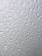 A collection of water droplets clings to a smooth, transparent surface, creating a pattern that is irregular but aesthetically pleasing. The droplets vary in size and are closely packed together, allowing light to pass through and create a reflective effect.