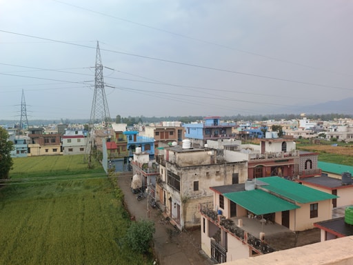 A small town or village with multiple houses, each having a variety of colors including blue, green, and beige. There are fields of green crops adjacent to the buildings. Power lines and large electrical pylons stretch across the skyline. The town appears to be surrounded by greenery, with hills visible in the distance under a cloudy sky.