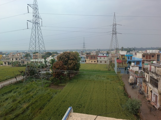 A rural landscape featuring a field of green crops with several tall electricity pylons extending into the distance. In the background, there are residential buildings with diverse architectural styles and colors, suggesting a developing area. The sky appears cloudy, and the scene is bordered by trees and a narrow road running alongside the fields.