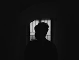 A shadowy silhouette of the corrupt officer standing against a barred prison window with red light casting ominous shadows.