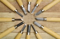A collection of wood carving tools arranged in a circular pattern on a wooden surface, featuring metal blades and light-colored wooden handles.