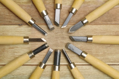 A collection of wood carving tools arranged in a circular pattern on a wooden surface, featuring metal blades and light-colored wooden handles.