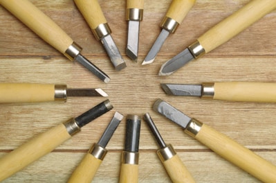 A collection of wood carving tools arranged in a circular pattern on a wooden surface, featuring metal blades and light-colored wooden handles.