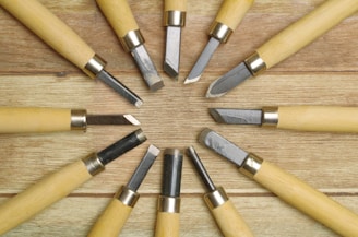 A collection of wood carving tools arranged in a circular pattern on a wooden surface, featuring metal blades and light-colored wooden handles.