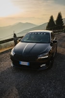 A black car is parked on a mountain road during sunset, surrounded by a scenic landscape of rolling hills and pine trees. The car features Japanese characters on the windshield.