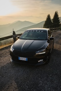 A black car is parked on a mountain road during sunset, surrounded by a scenic landscape of rolling hills and pine trees. The car features Japanese characters on the windshield.