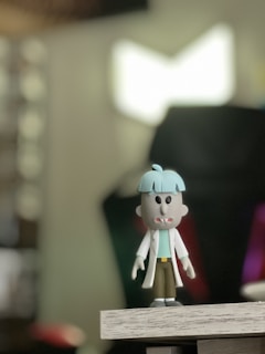 A cartoonish toy figure stands on a wooden surface. The figure has a distinctive light blue bowl-shaped haircut and wide eyes with a small, open mouth showing two fangs. It is dressed in a white coat over a green shirt and brown pants. The background is blurry, displaying a mix of soft colors and shapes, possibly indicating an indoor setting.