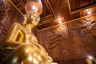A large golden statue of Buddha is prominently displayed in the room, surrounded by intricately carved wooden walls depicting various scenes and patterns. The ceiling features wood paneling with embedded lights, and the atmosphere exudes an aura of grandeur and peace.