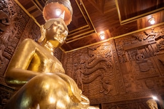 A large golden statue of Buddha is prominently displayed in the room, surrounded by intricately carved wooden walls depicting various scenes and patterns. The ceiling features wood paneling with embedded lights, and the atmosphere exudes an aura of grandeur and peace.