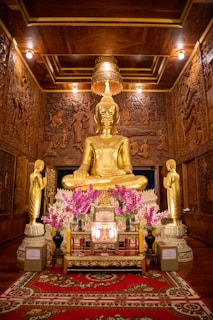 A golden Buddha statue is seated on an ornate platform in a room with intricately carved wooden walls. Flanking the central statue are two smaller standing figures. Flower arrangements with purple blossoms are placed around the statues. The ceiling is richly decorated with woodwork, and a golden chandelier hangs above.